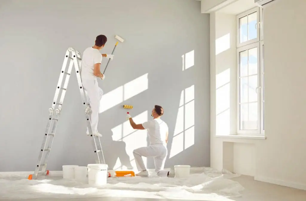 Two professional painters rolling gray paint on an interior wall, showing how crew size affects how long it takes to paint the interior of a house.