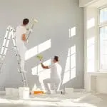 Two professional painters rolling gray paint on an interior wall, showing how crew size affects how long it takes to paint the interior of a house.